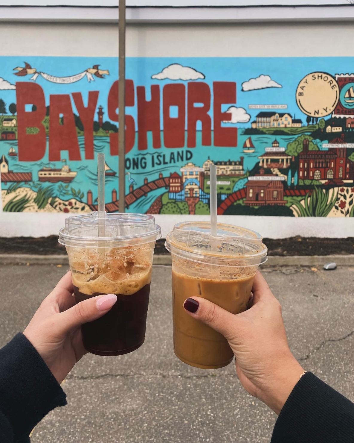 Two iced coffees held up in front of the Greetings from Bay Shore mural